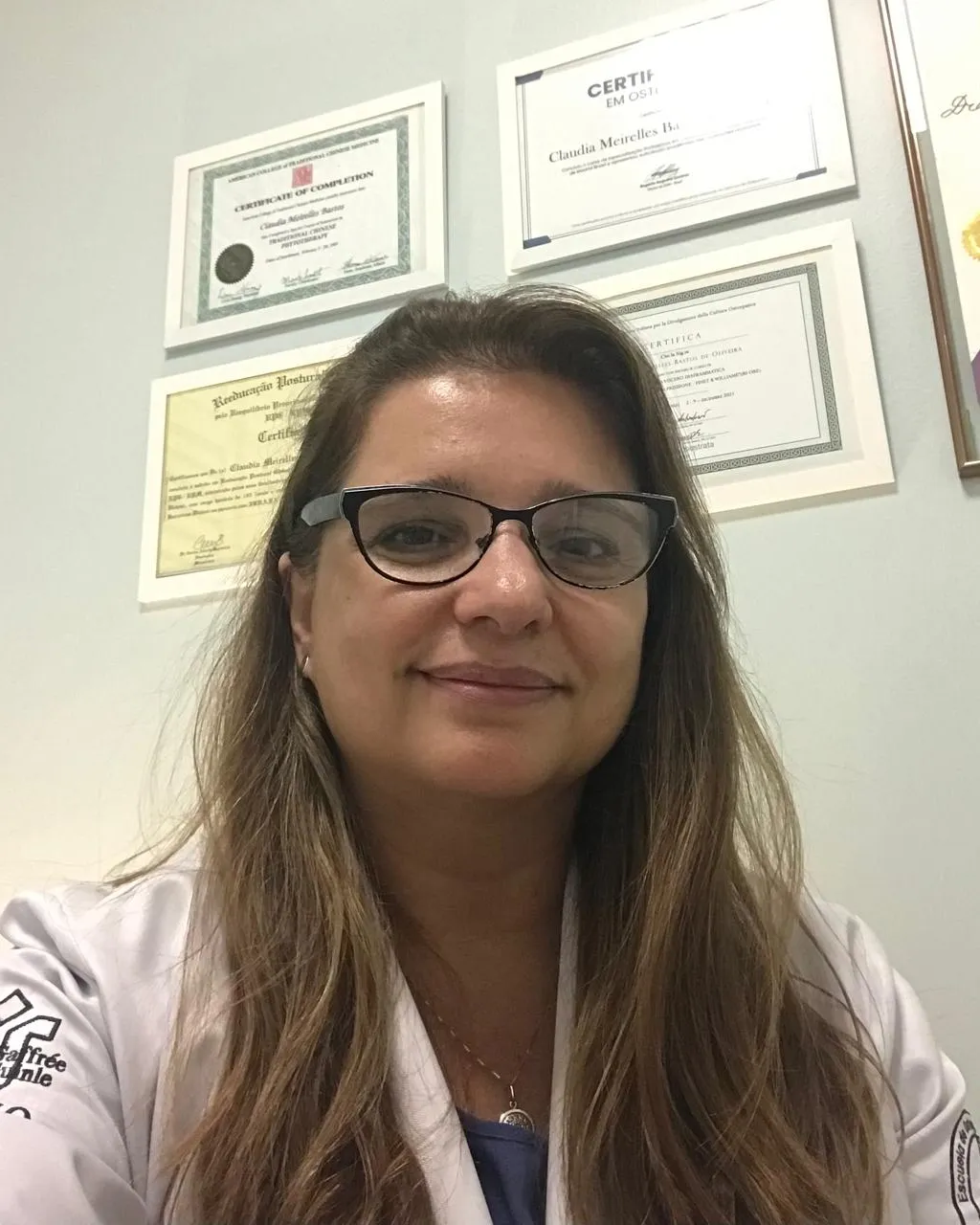 Dra. Claudia Meirelles, de jaleco branco, no consultório; atrás dela, os certificados de formação emoldurados na parede.